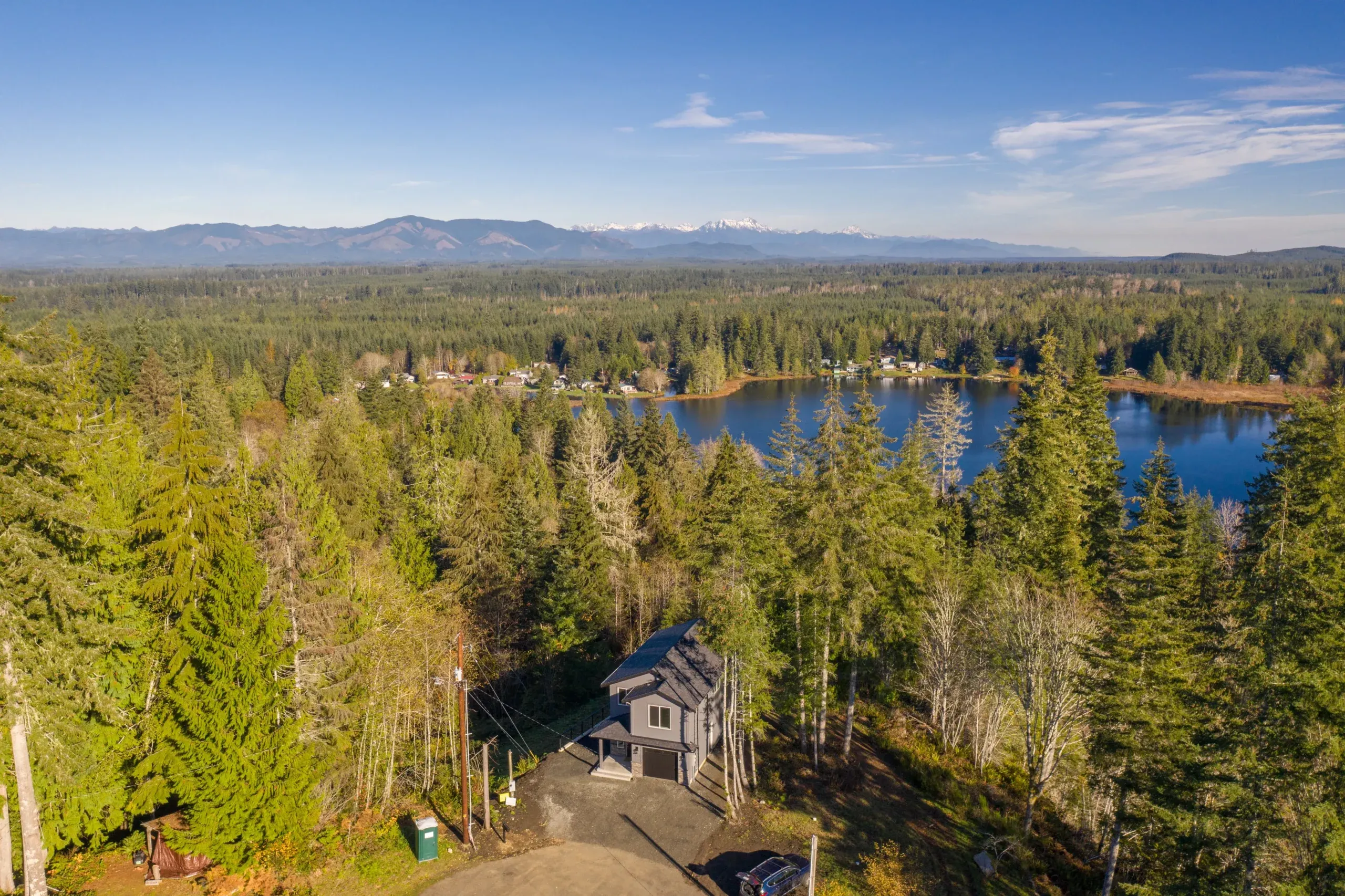 Aerial view of a forested lakeside with a small house and distant mountains under a clear sky