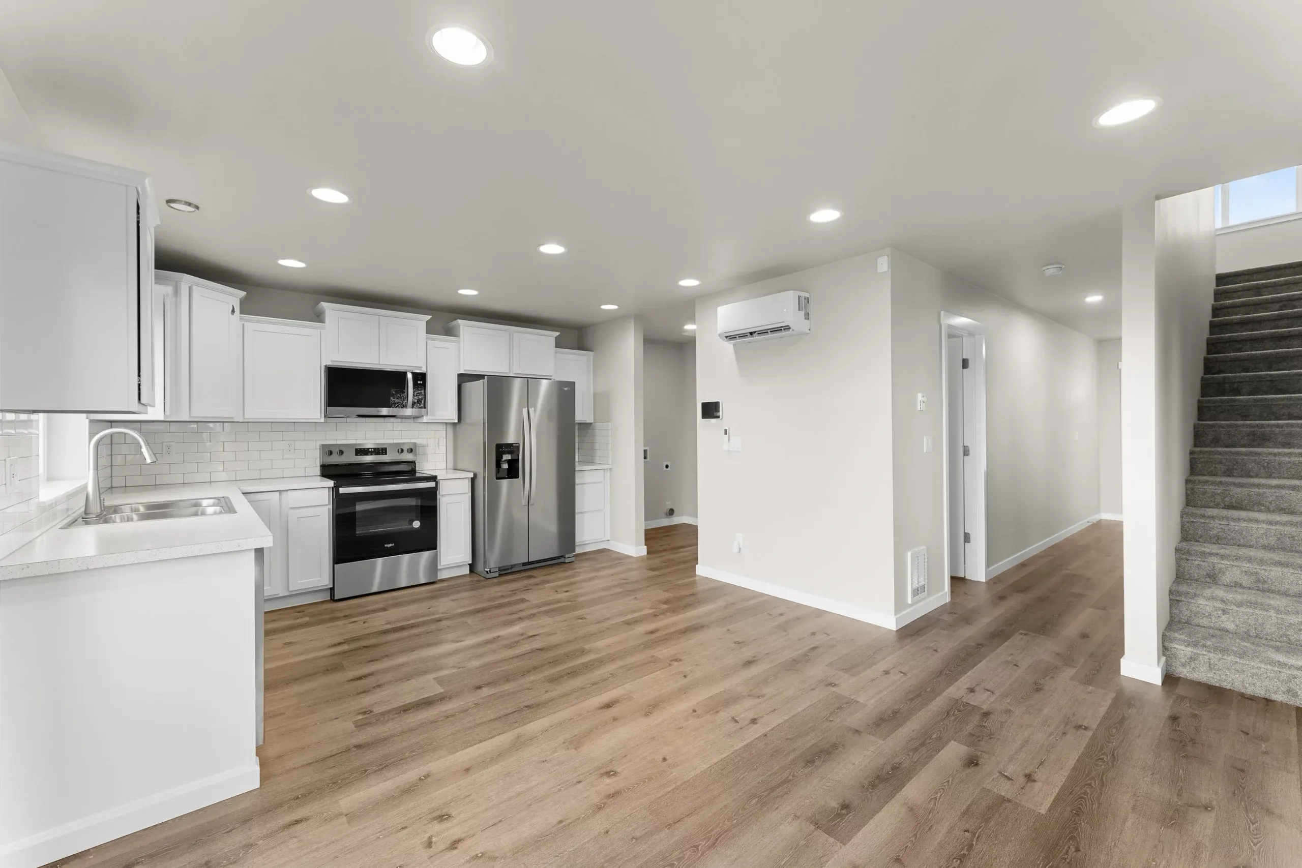 Bright modern open-plan kitchen and living space with white cabinets, stainless appliances, wood floors, and stairs.