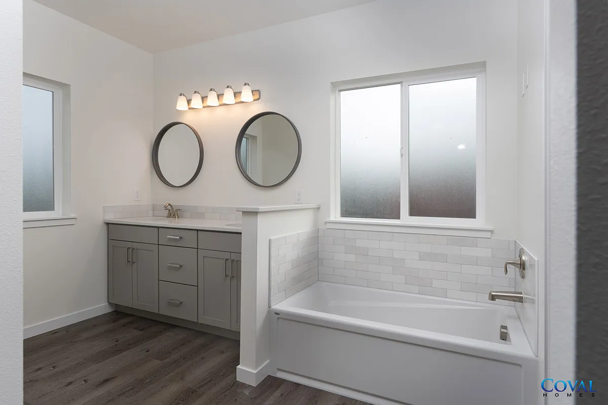 Bright white bathroom with double vanity, round mirrors, tub, and frosted window.