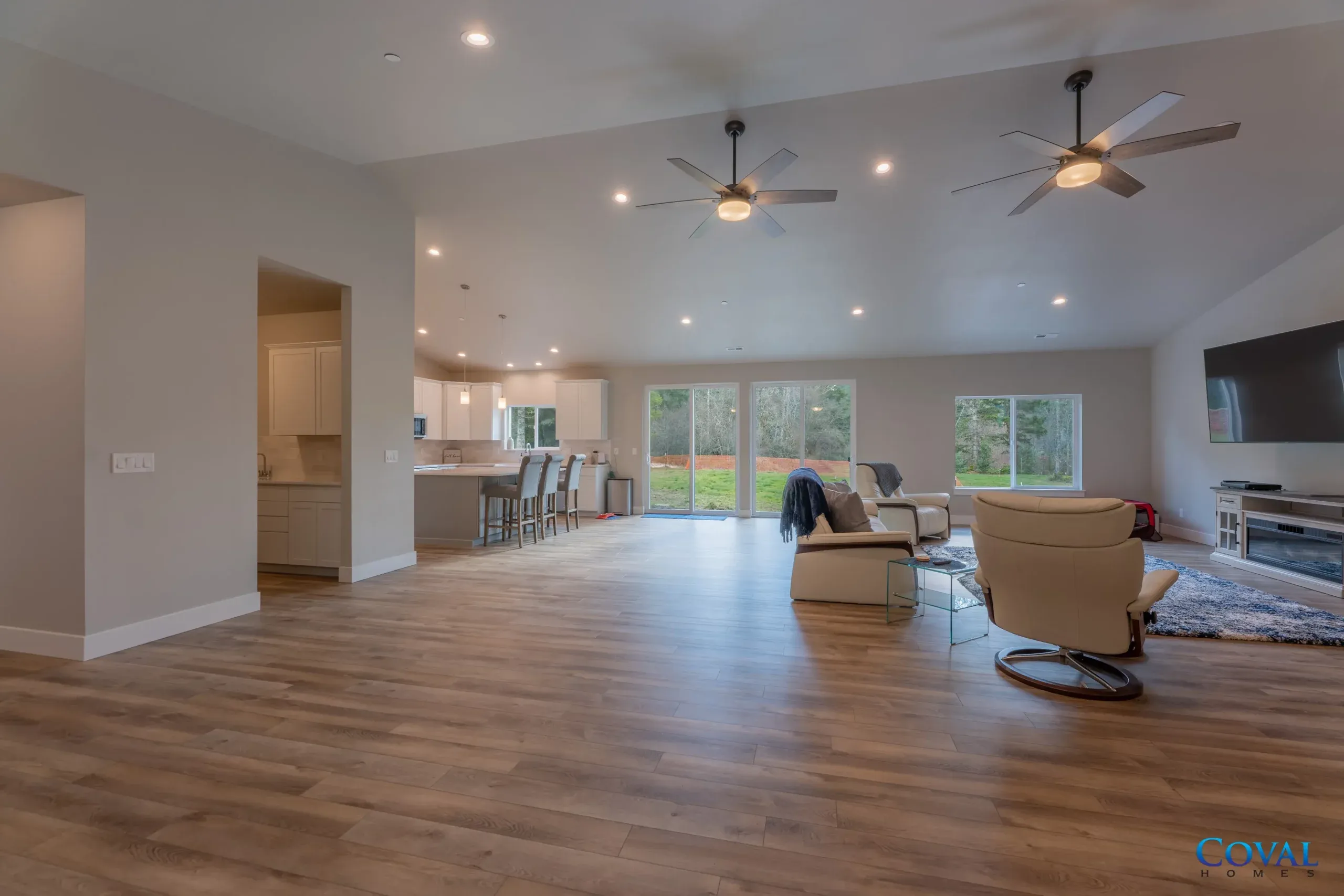 Bright open-concept living room with wood floors, ceiling fans, and large windows overlooking a backyard.
