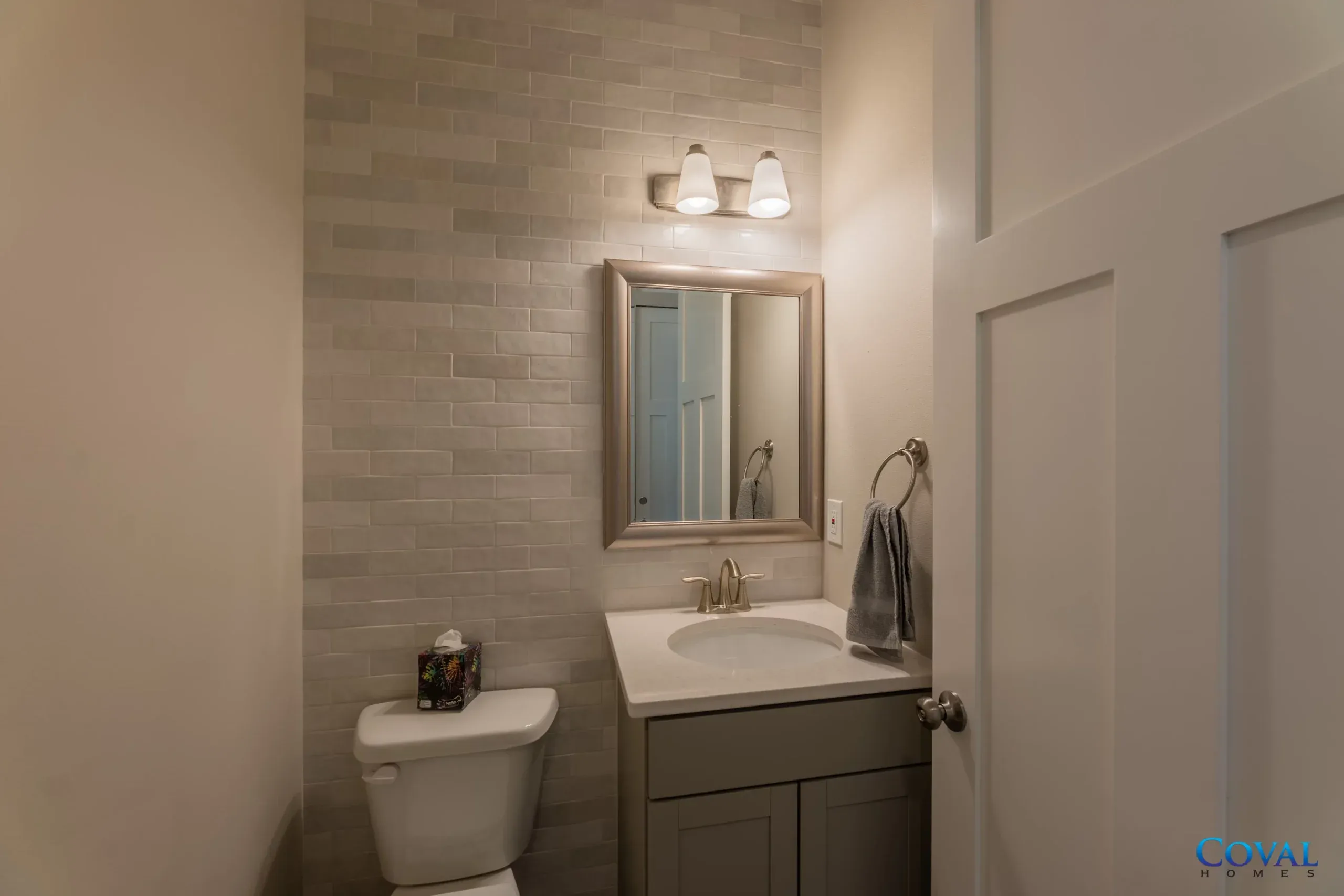 Small bathroom with white sink, mirror, toilet, and gray vanity against a light brick wall.