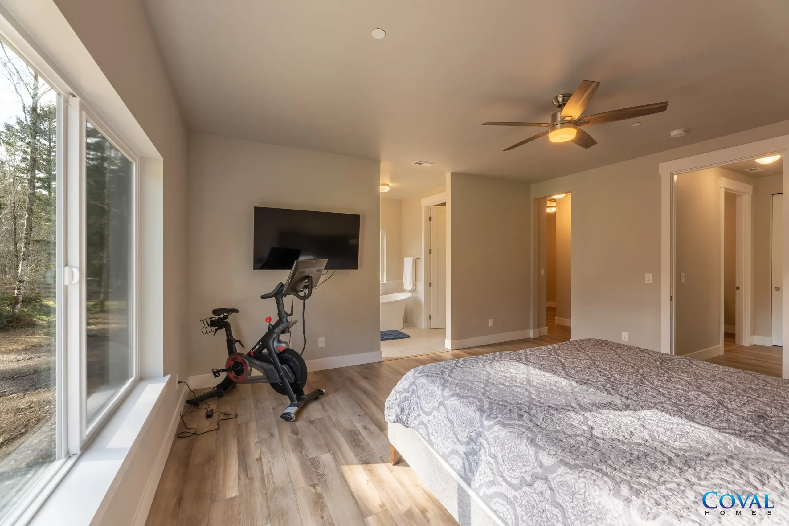 Bedroom with large windows, wall-mounted TV, exercise bike, and neutral decor with a ceiling fan