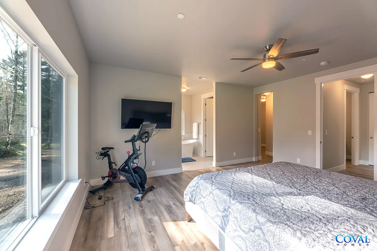 Bright bedroom with large windows, ceiling fan, wall TV, exercise bike, and a bed by the far wall.