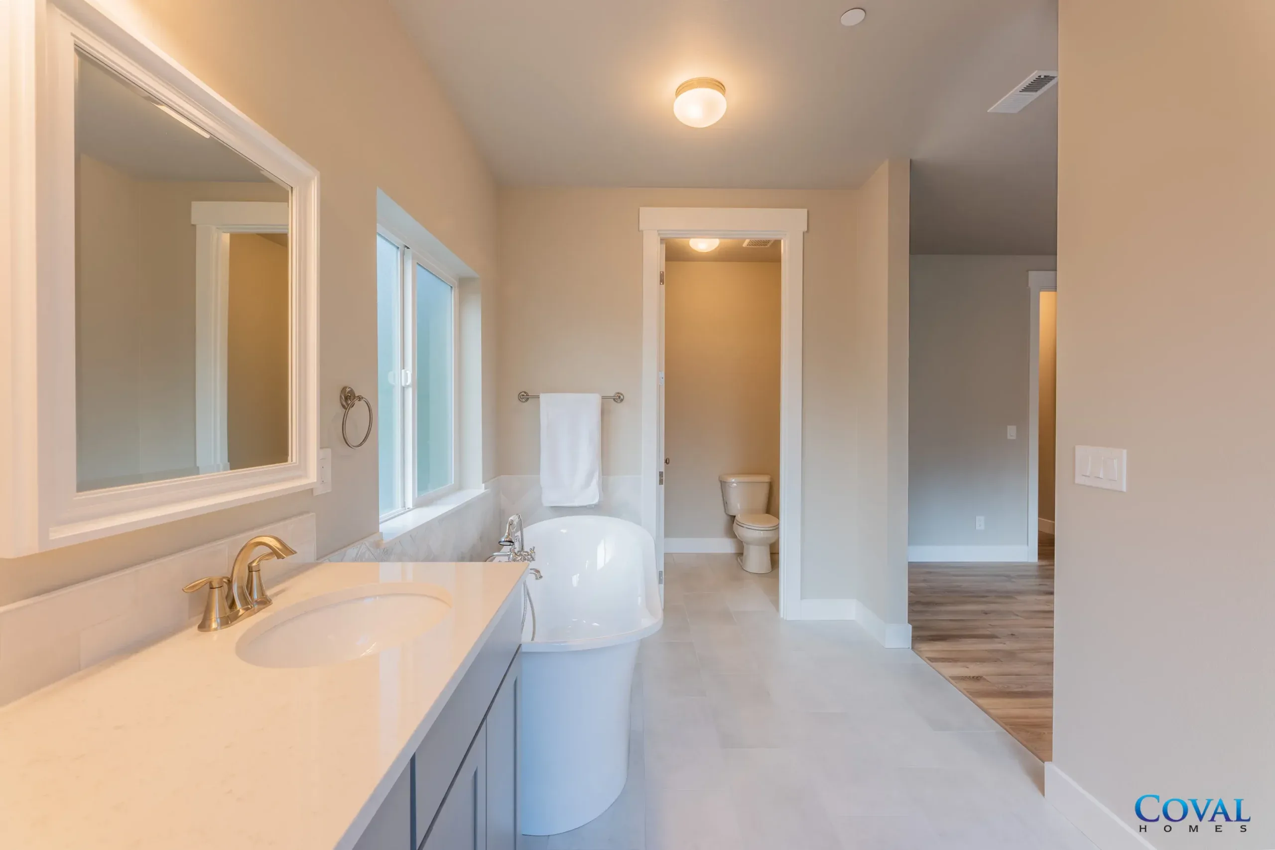 Bright bathroom with vanity, mirror, toilet, and tiled floor leading to an adjacent room.