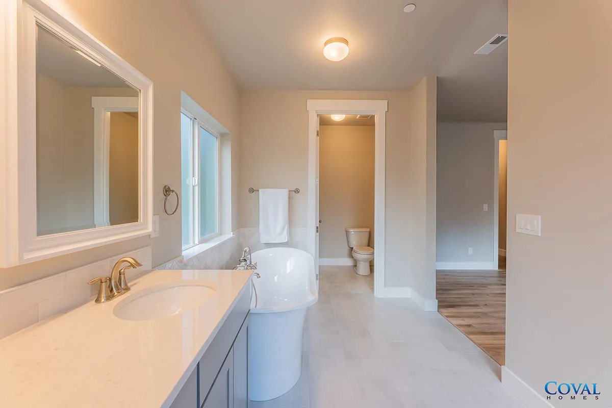 Bright beige bathroom with double vanity, mirror, toilet, and tiled floor leading to an adjoining room.