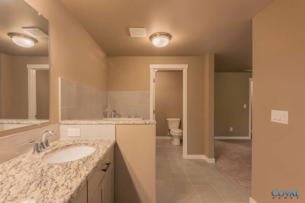 Bathroom with double vanity, beige tile, and a separate toilet room visible in the background