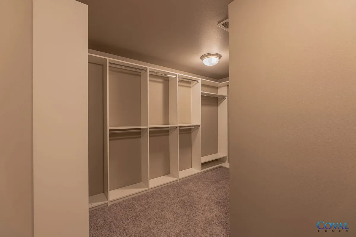 Empty carpeted walk-in closet with white built-in shelves and hanging rods, lit by a ceiling fixture