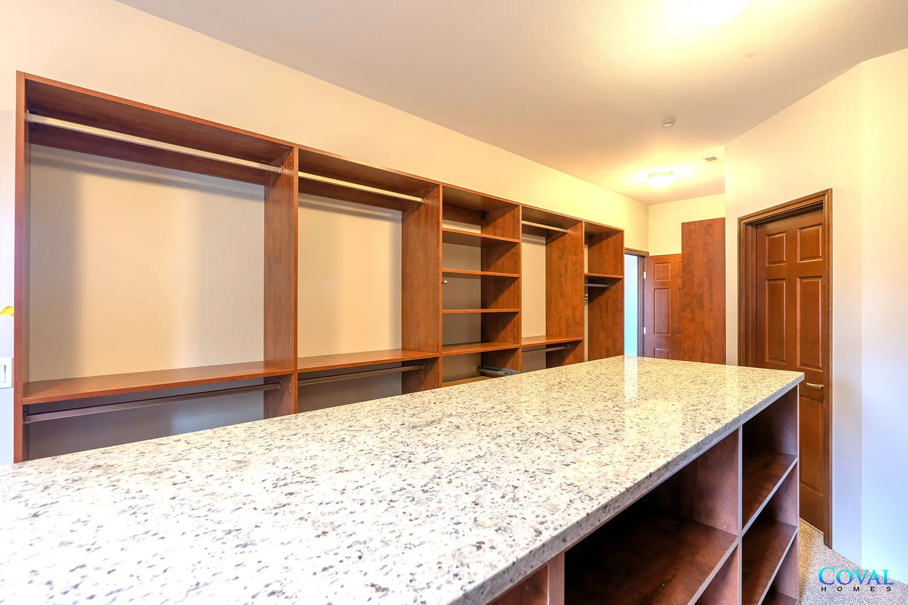 Empty walk-in closet with wooden shelves, hanging rods, and a speckled countertop under warm ceiling lights