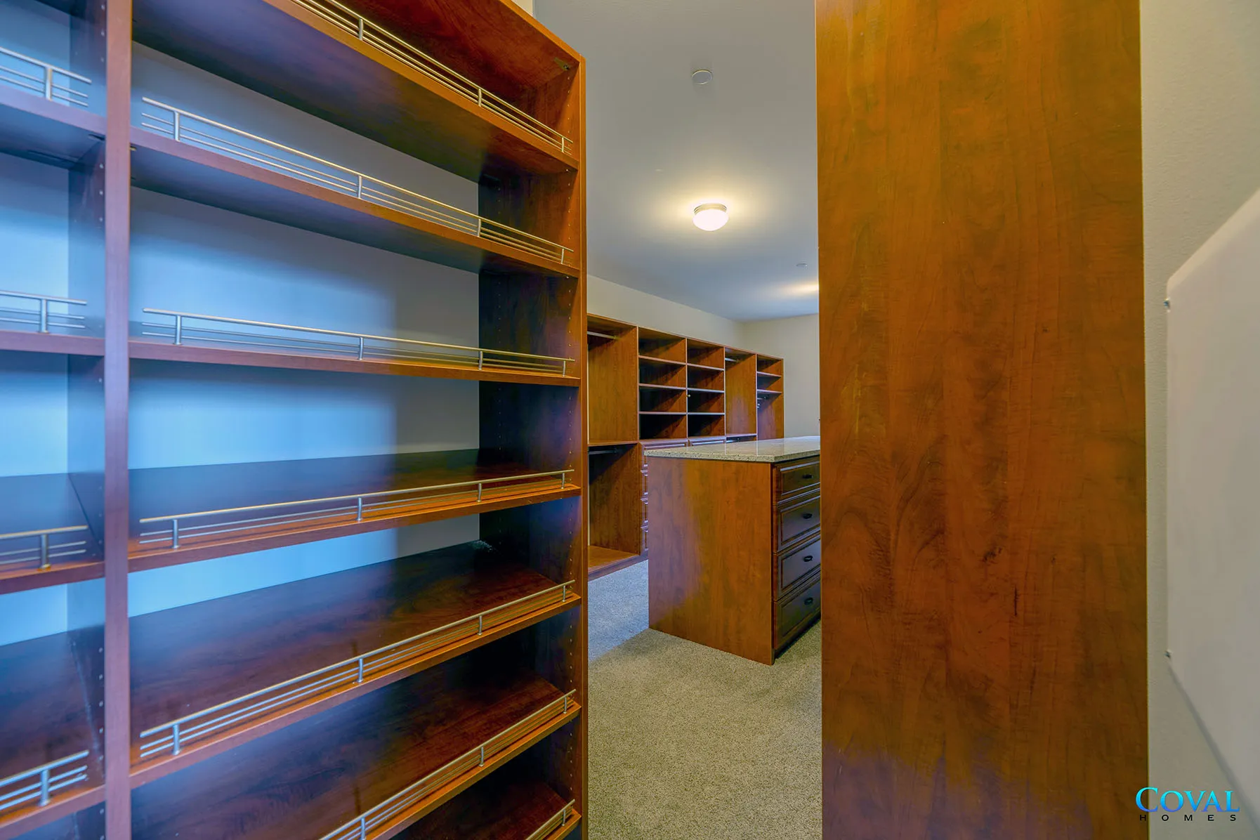 Small walk-in closet with wooden shelves, carpeted floor, and a narrow hallway view to more shelving.