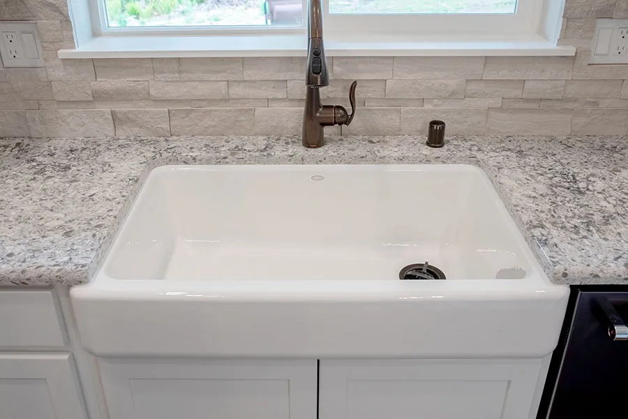 White farmhouse sink with black faucet beneath a window, set in a light granite countertop.