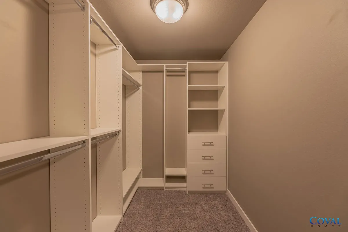 Empty walk-in closet with white shelves, hanging rods, drawers, and beige walls under a ceiling light.