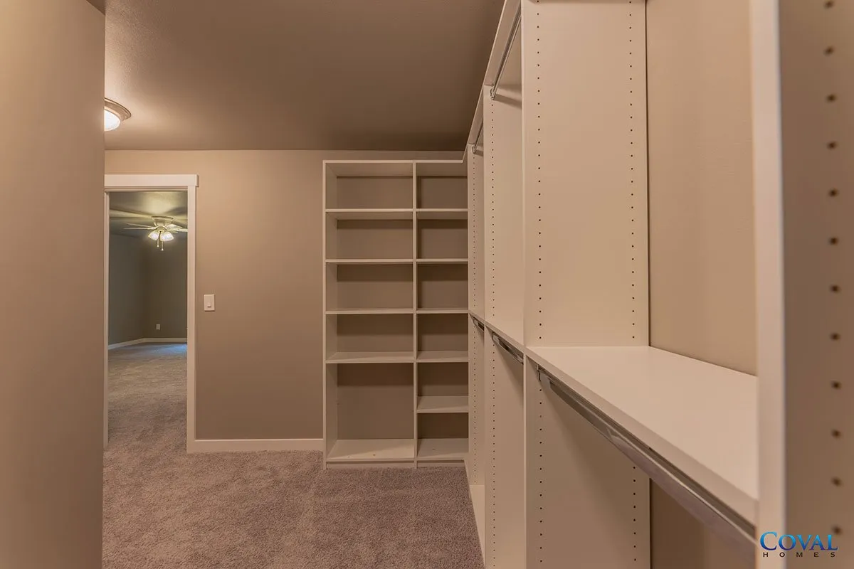 Empty walk-in closet with beige walls, white built-in shelving, and carpeted floor leading to a doorway.