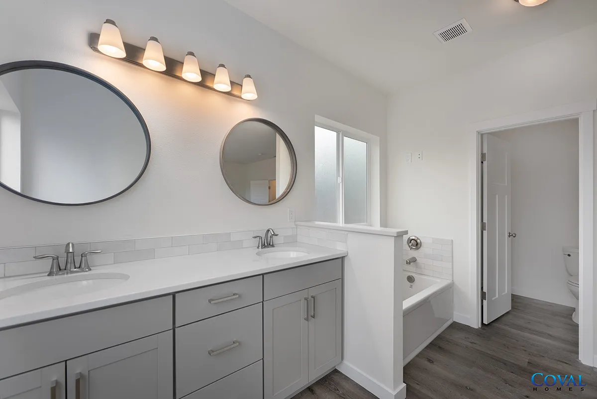 Bright white bathroom with double sinks, round mirrors, and gray vanity cabinets