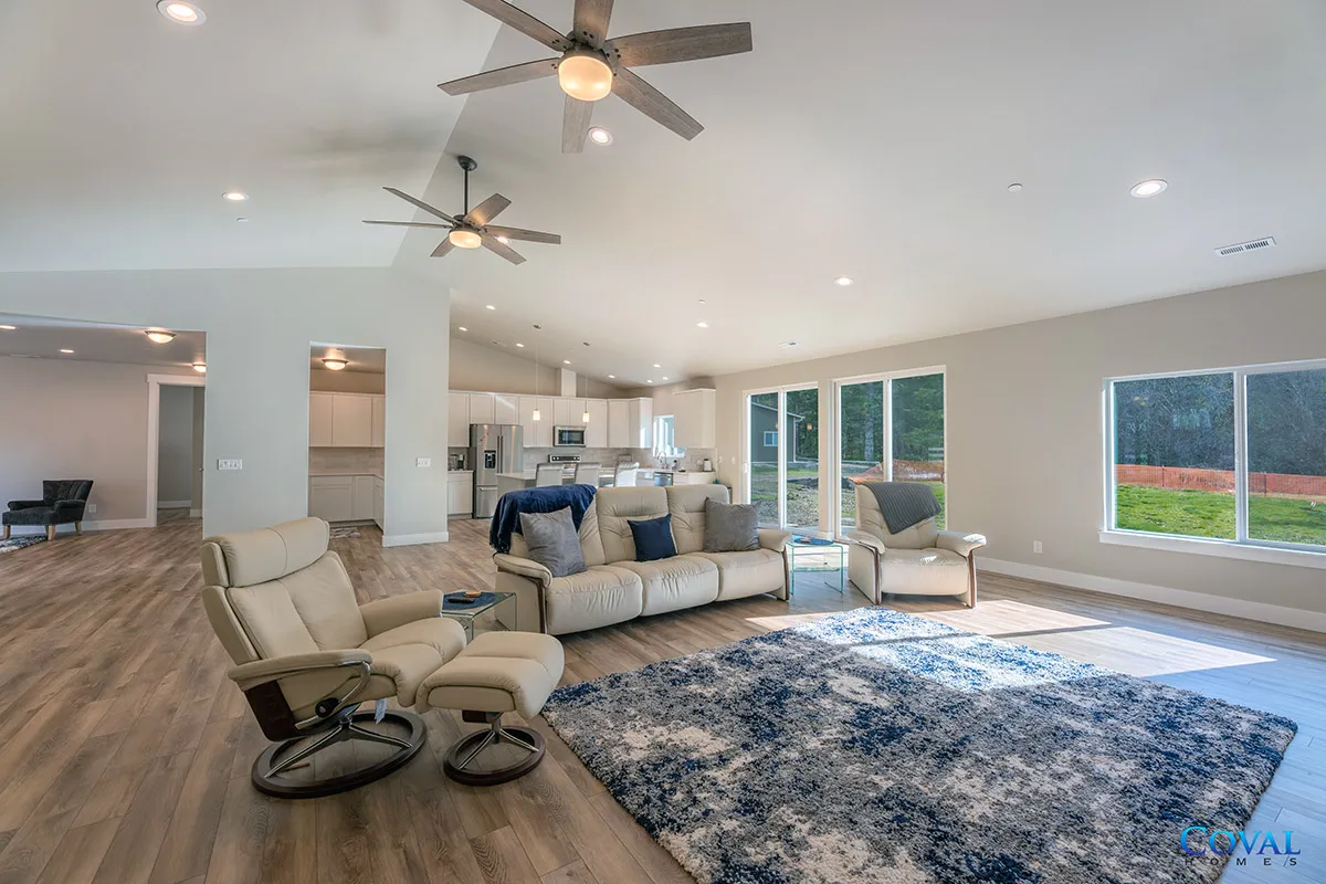Bright living room with recliners, sectional sofa, blue rug, ceiling fans, and large windows overlooking a yard