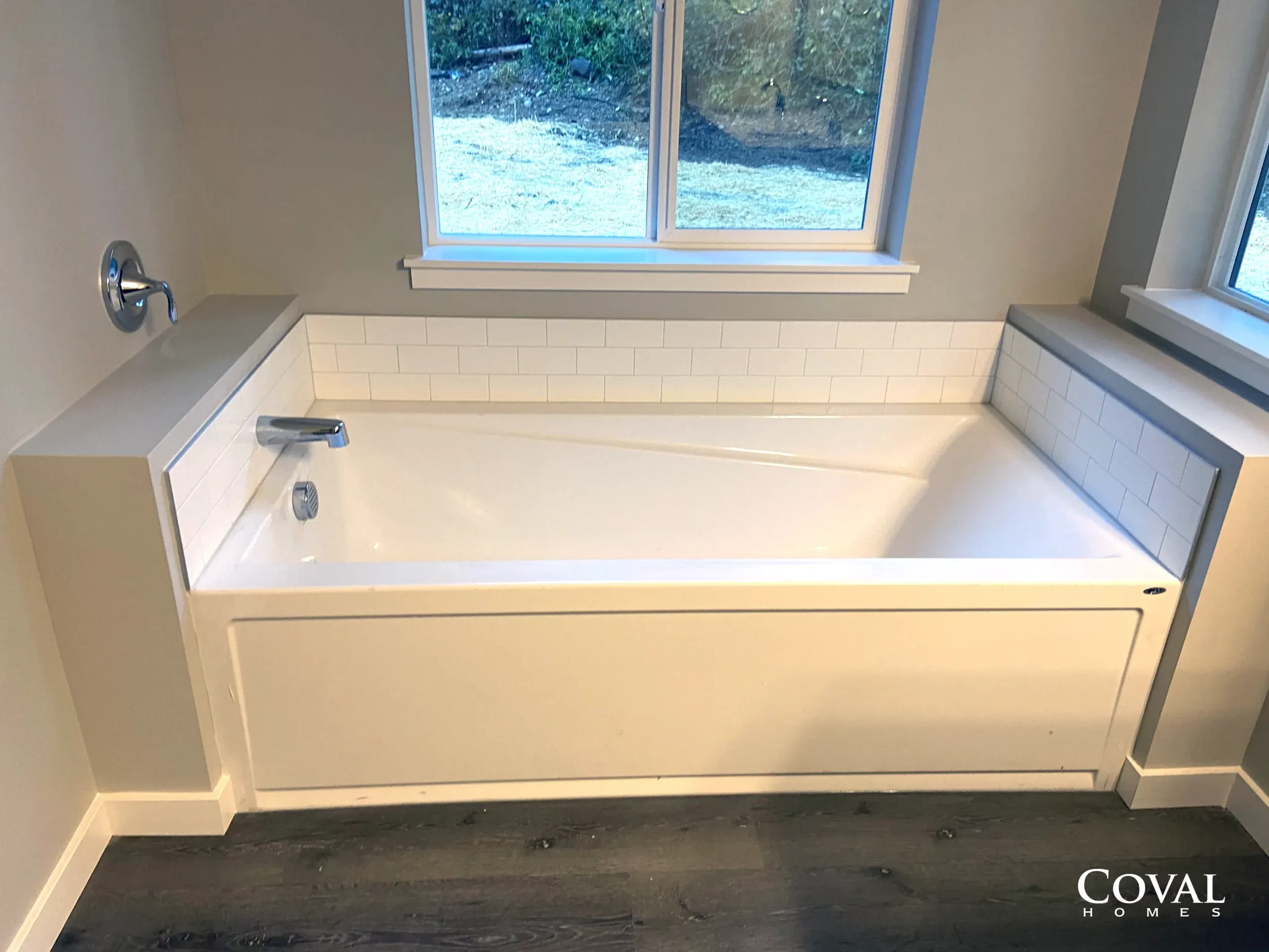 Bathroom with a white soaking tub beneath a window and tiled surround, in a gray room.