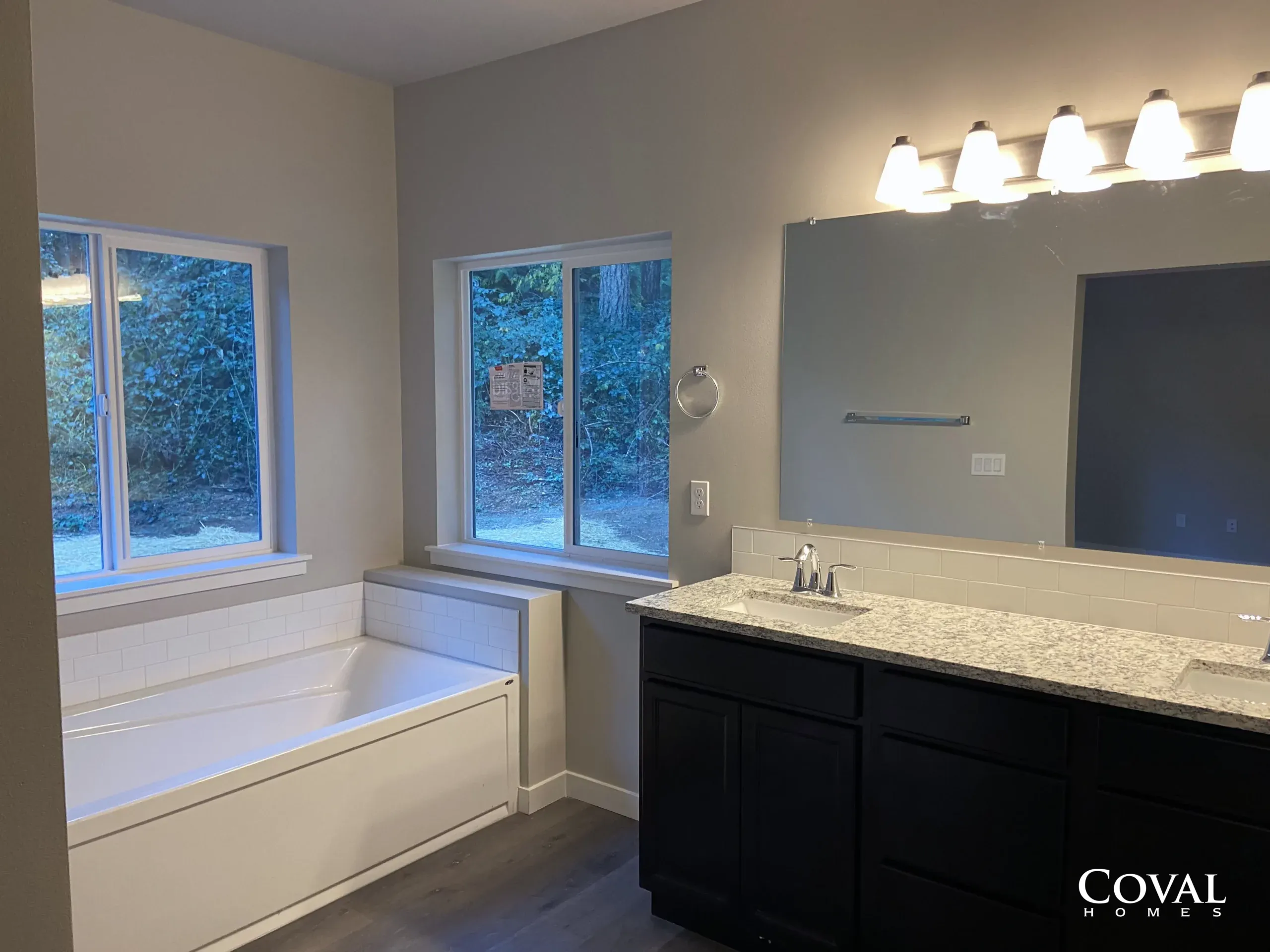 Bathroom vanity and bathtub area with double sinks, large mirror, and windows showing trees outside