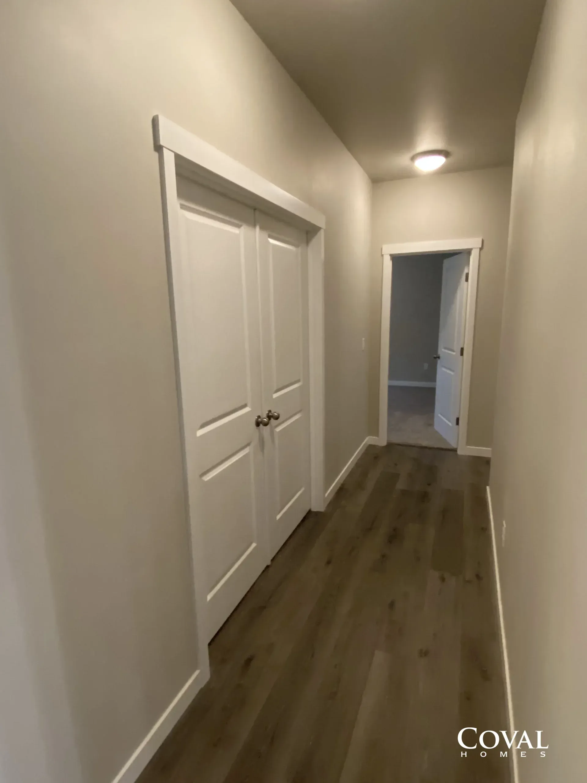 Narrow hallway with white doors, wood floor, and ceiling light leading to a bathroom.