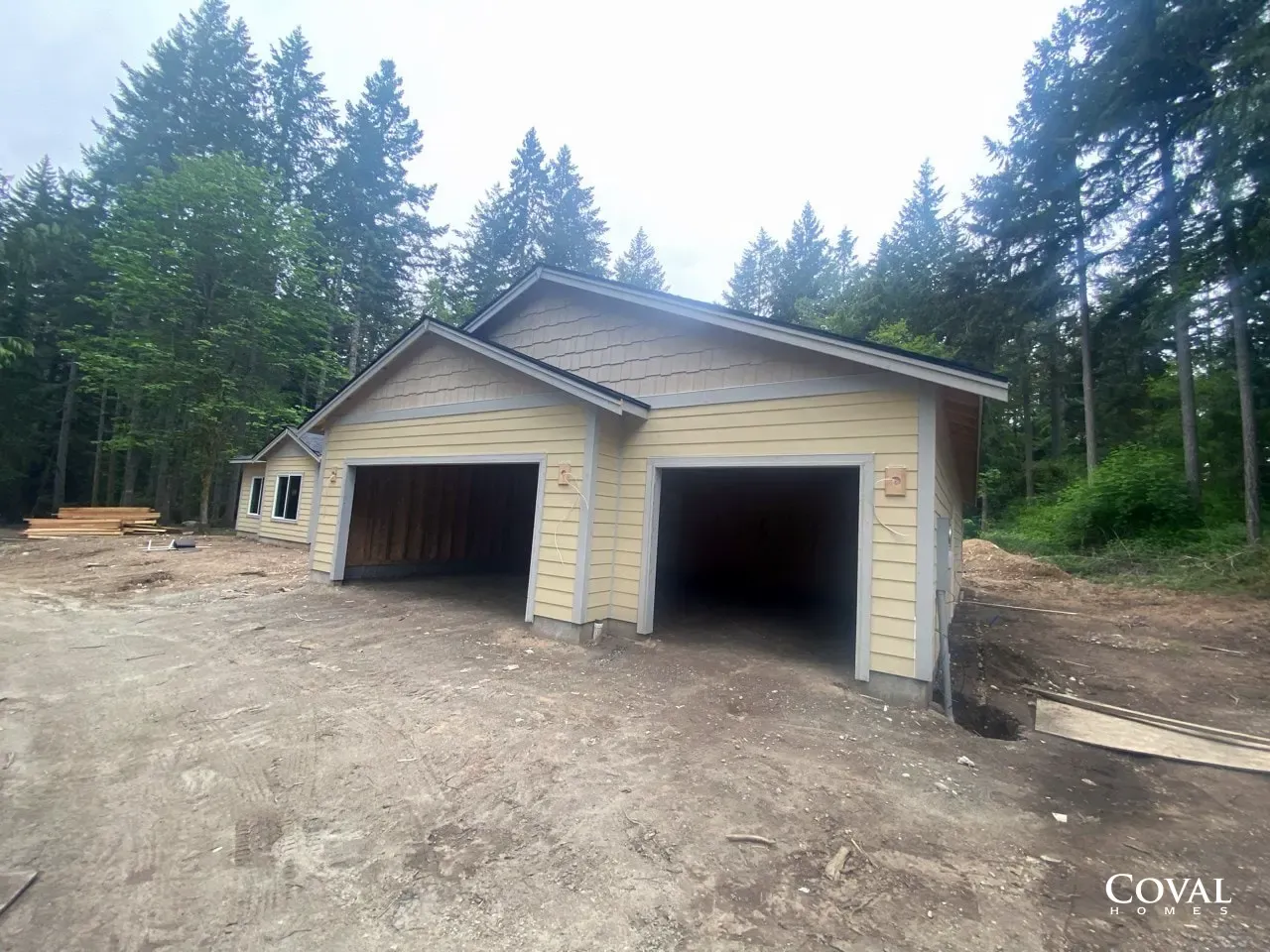 Two-car garage with beige siding on a dirt lot, surrounded by trees and forest.