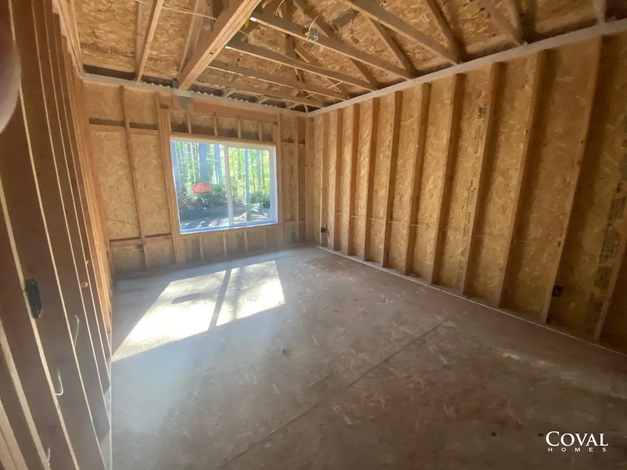 Unfinished room with exposed wooden framing, plywood walls, and sunlight through a large window.