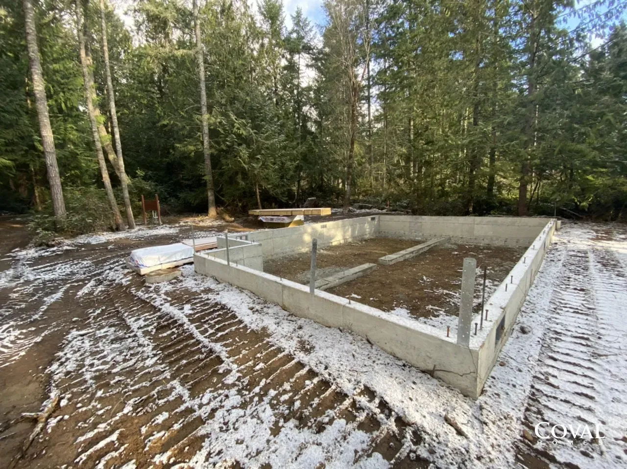 Concrete foundation in a snowy wooded clearing, with partial walls and nearby materials.