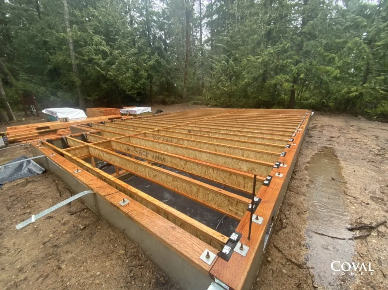 Wooden deck framing under construction on a dirt lot beside trees, with joists and beams installed.