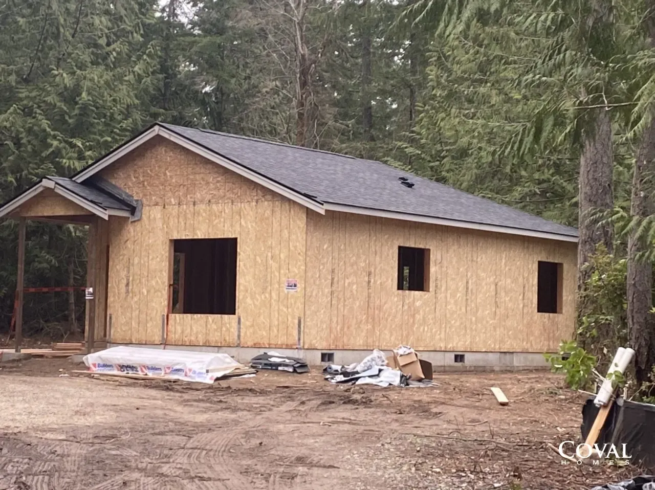 Small unfinished wooden house under construction in a forest clearing with exposed framing and open windows