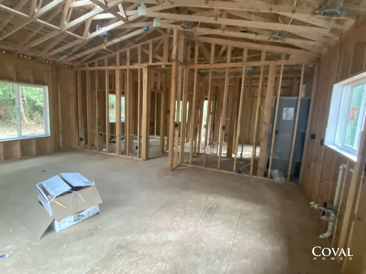 Interior of a framed house under construction with exposed wood studs and trusses, unfinished floor, and windows.