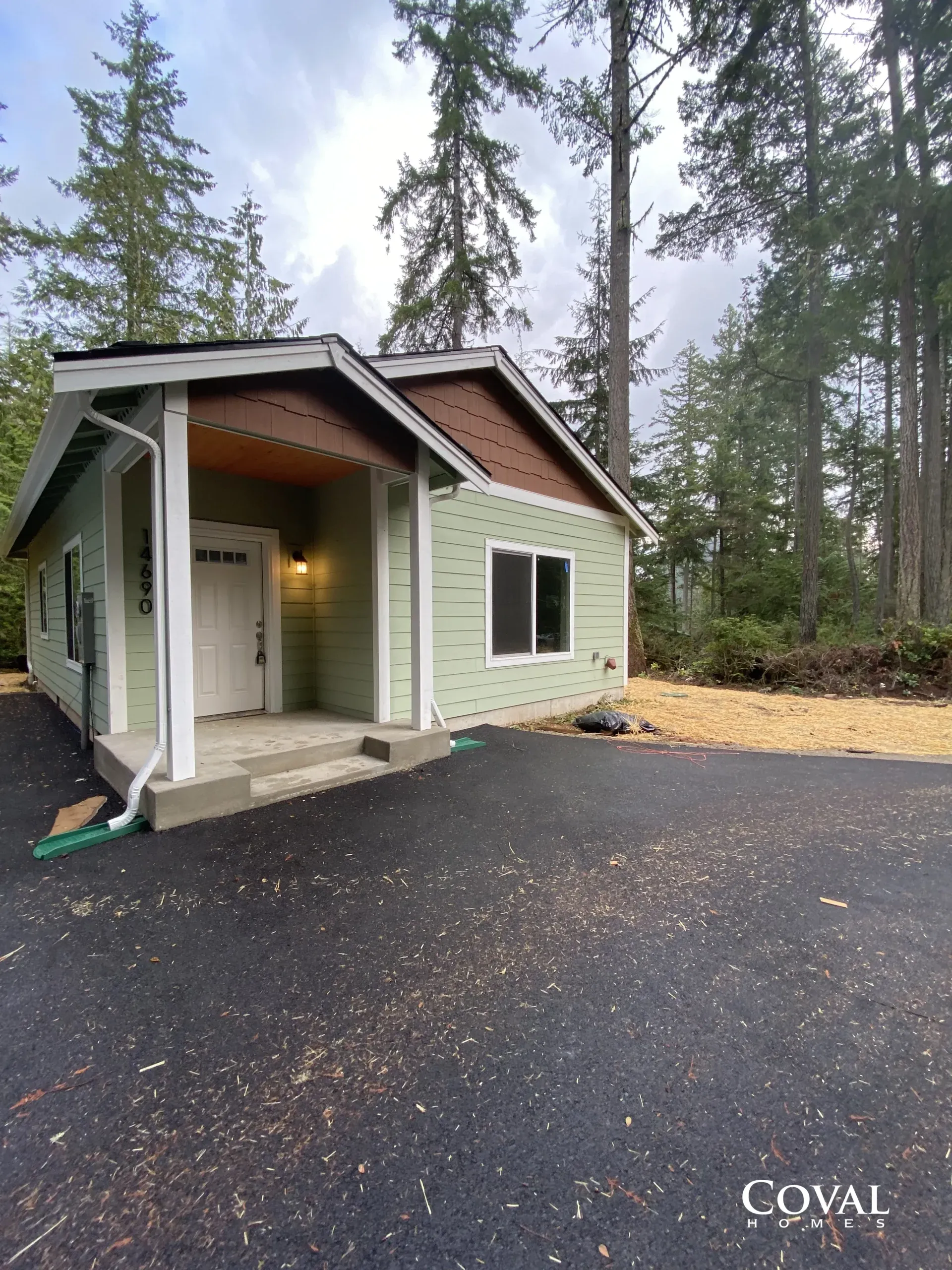 Small green cabin with covered porch beside a wet asphalt driveway in a wooded area.