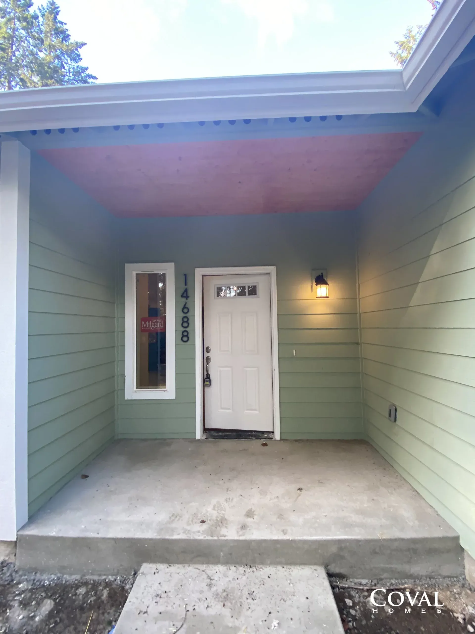 Front porch with white door, sage-green siding, wall light, and concrete steps leading up to the entrance.