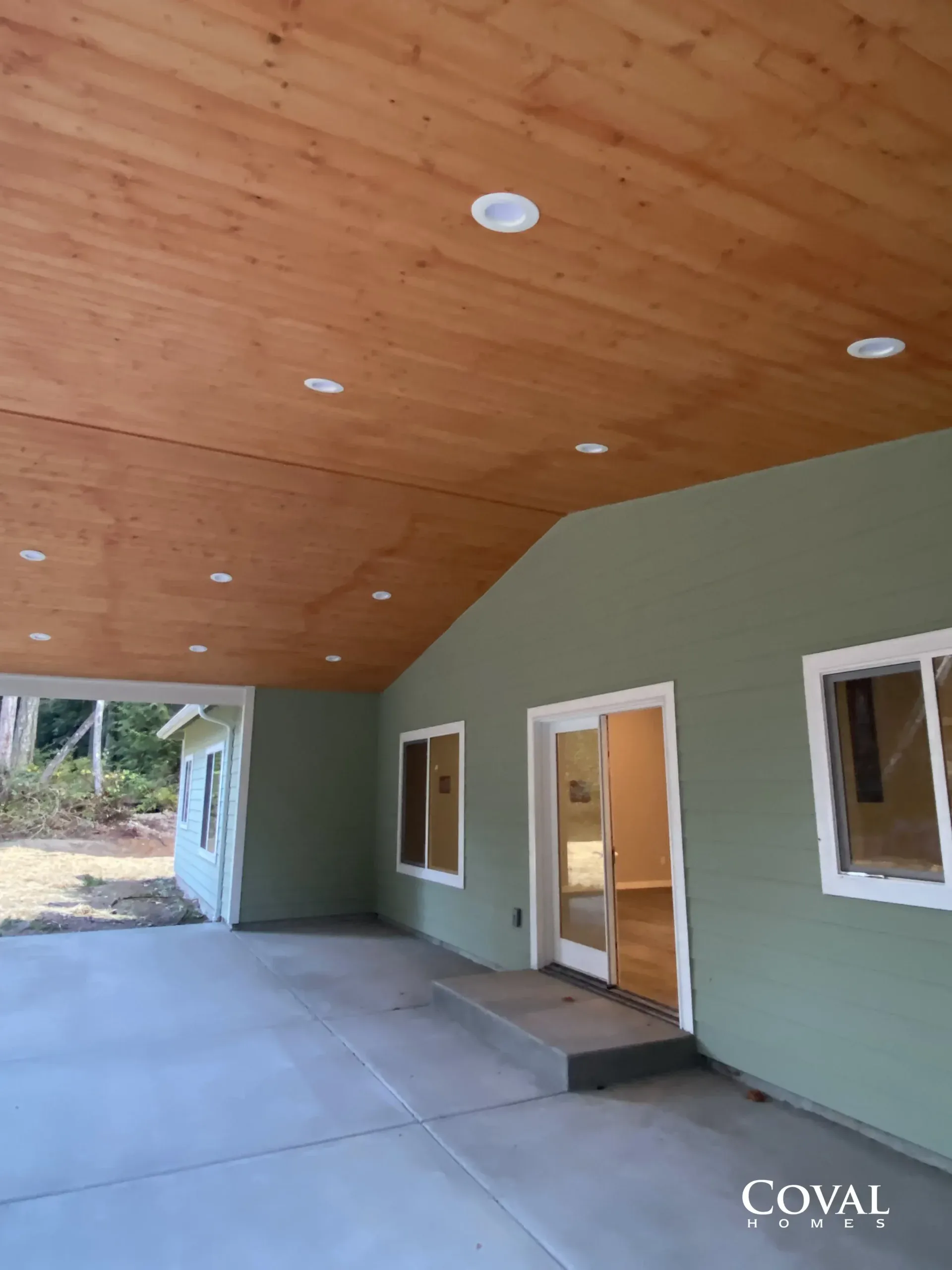 Covered porch with wood ceiling, green walls, windows, and open doorways beside a concrete patio.