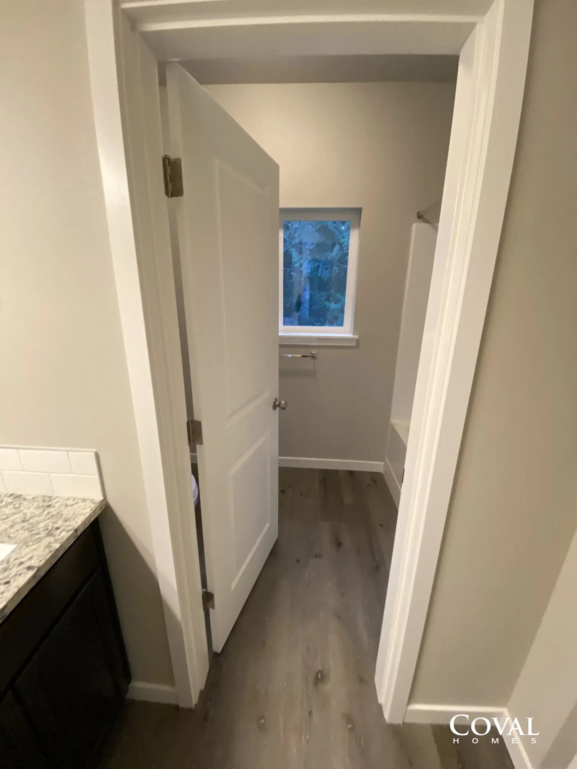 Narrow hallway with open white doors, gray wood floor, and a small window at the end.