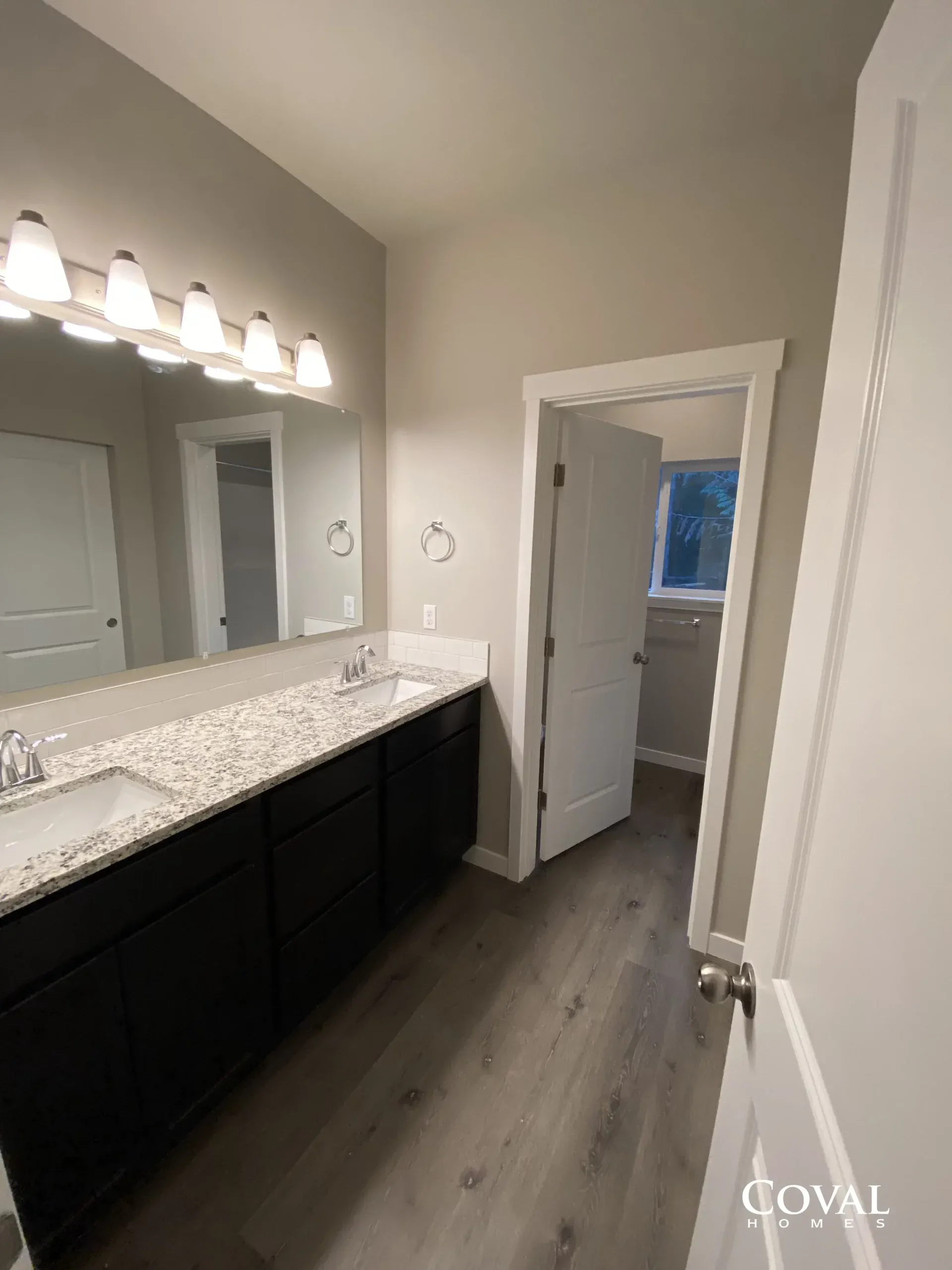 Modern bathroom with double vanity, large mirror, white countertop, and dark cabinets, viewed toward a doorway