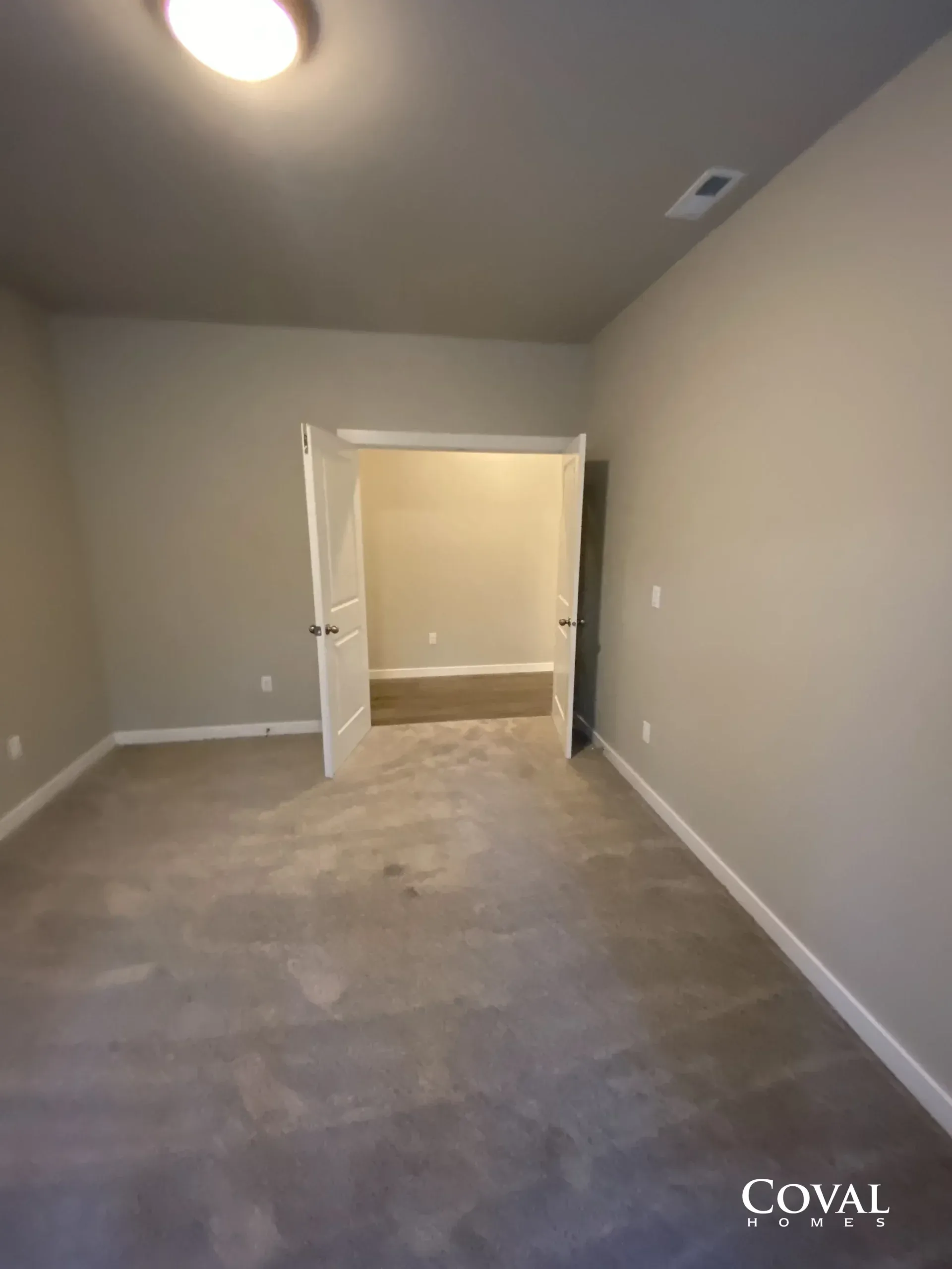 Empty beige room with gray carpet, ceiling light, and open doorway to a small adjoining space.