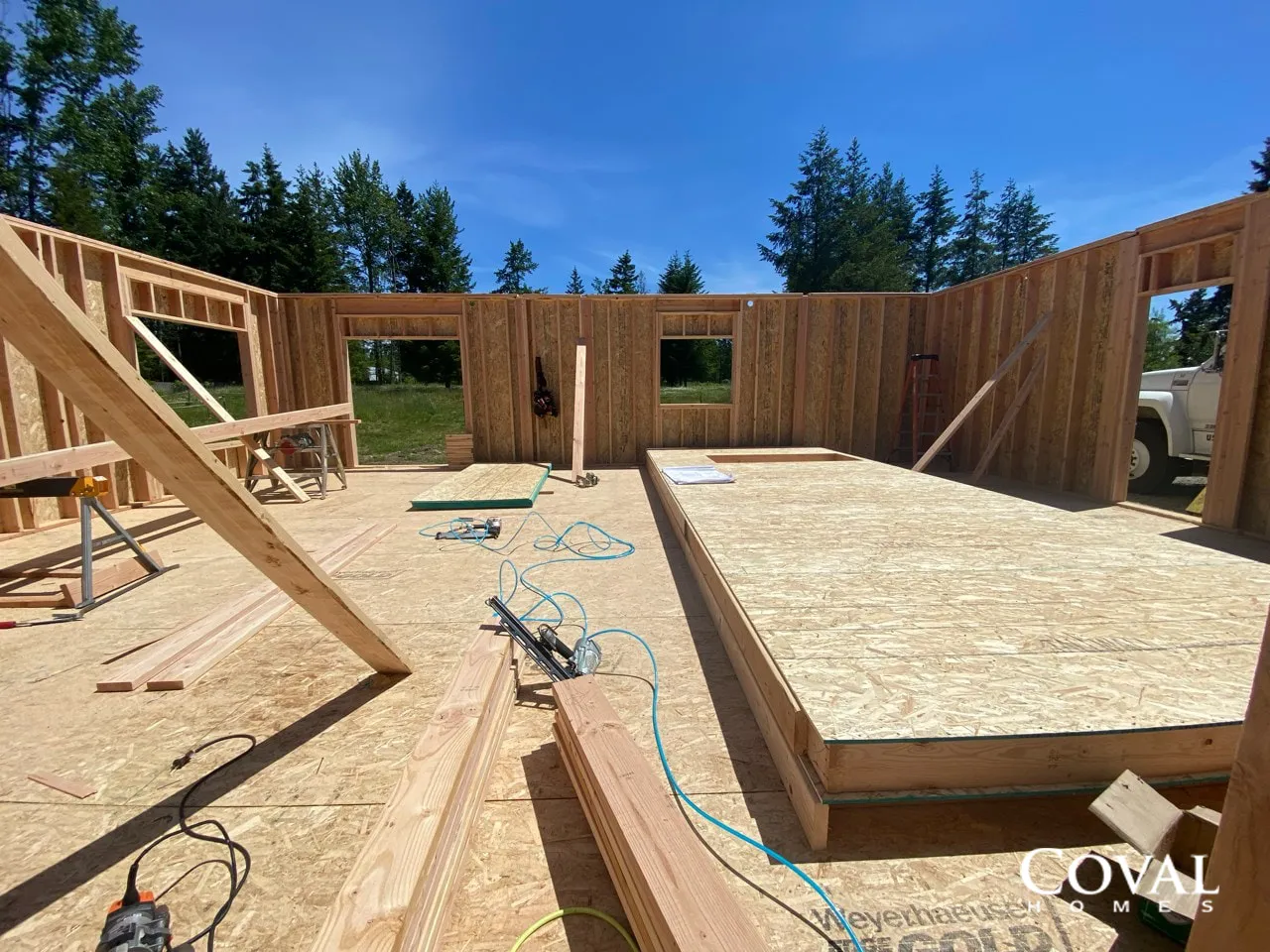 Wood-framed house under construction, with plywood subfloor and wall framing in a sunny open interior.