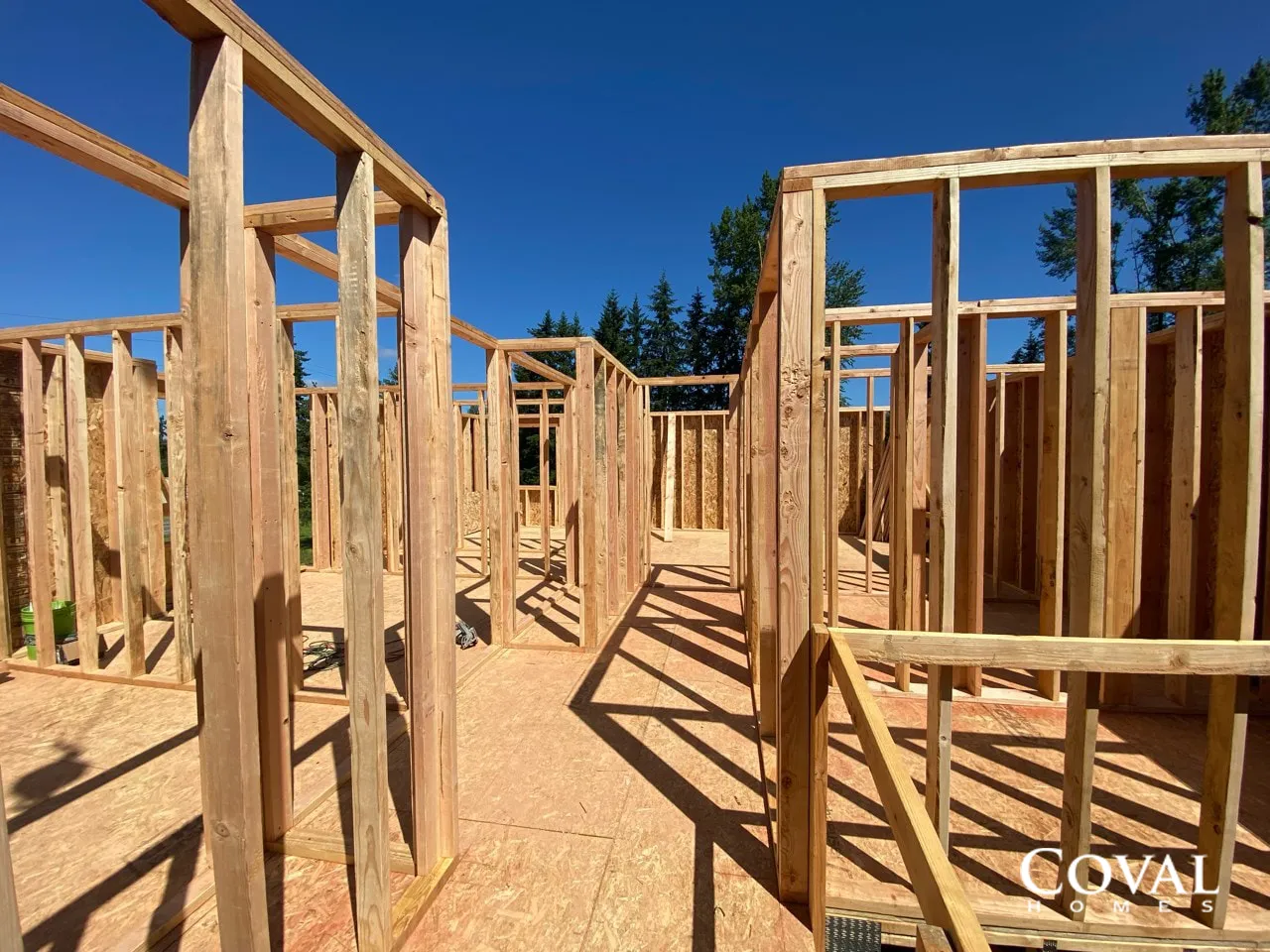 Wood-framed house under construction against a clear blue sky, with a sandy ground and shadows.