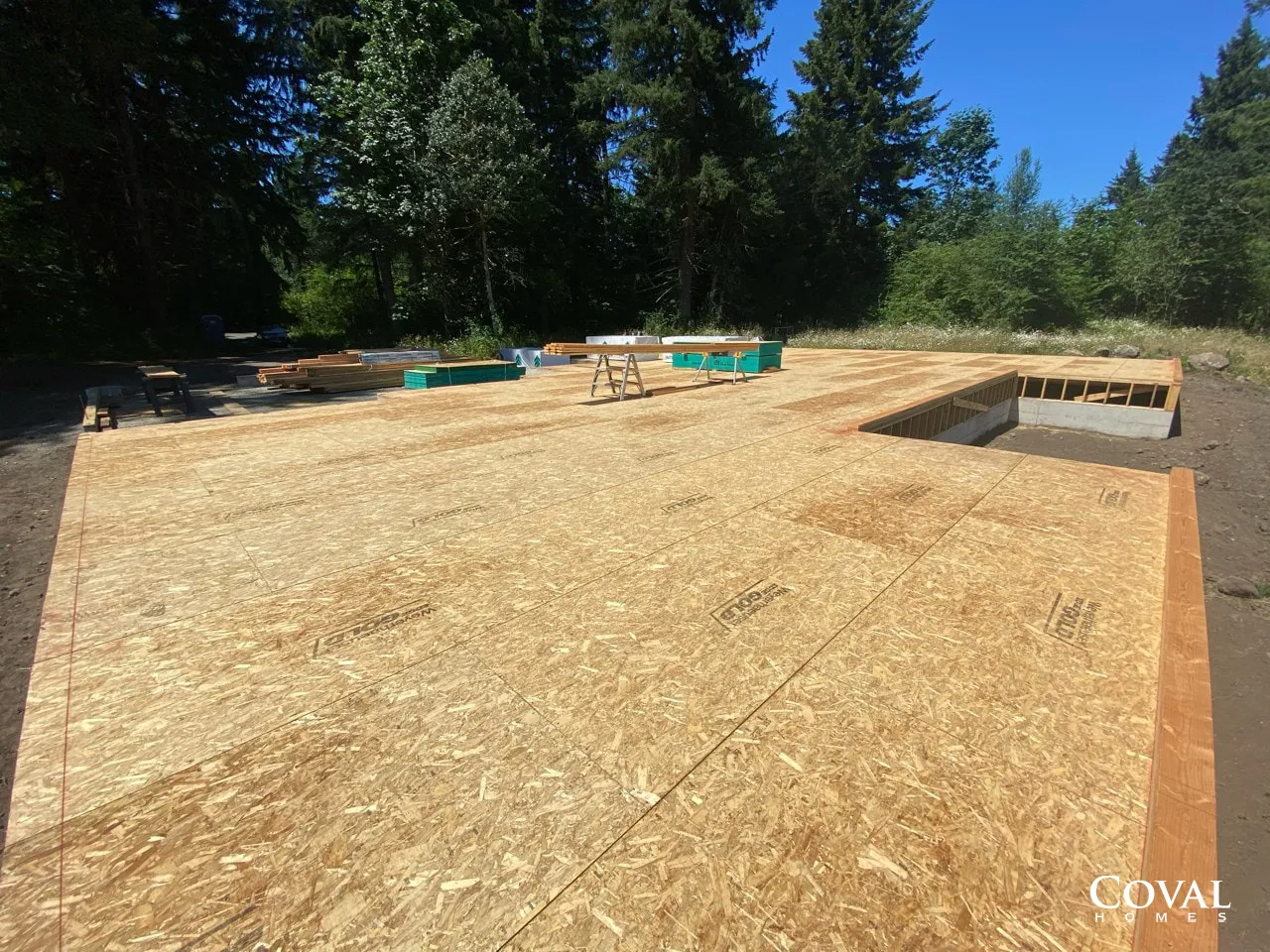 New wood subfloor under construction beside a framed rectangular opening, surrounded by trees under a blue sky