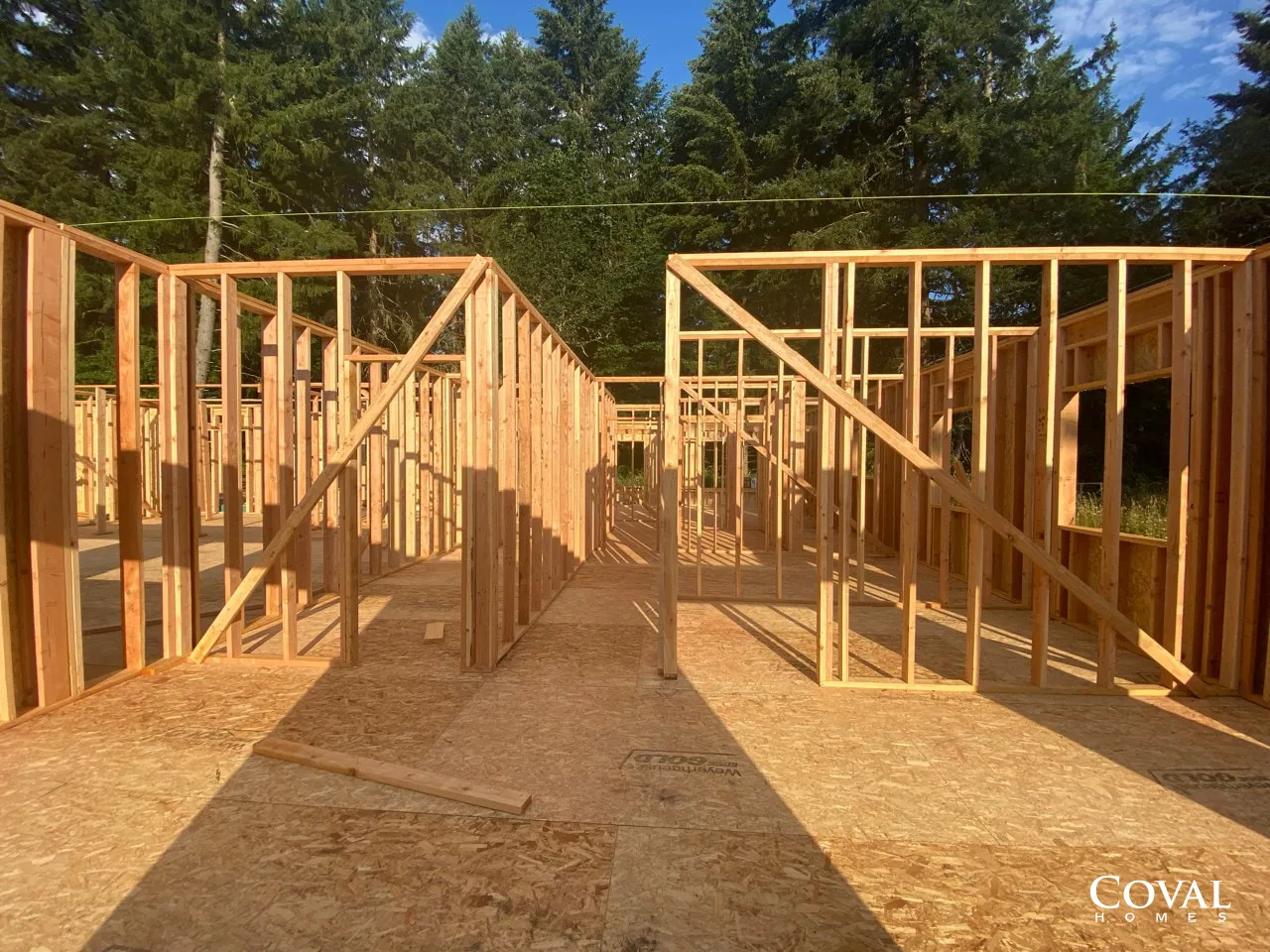 Wooden house framing under construction on a sunny lot with trees in the background