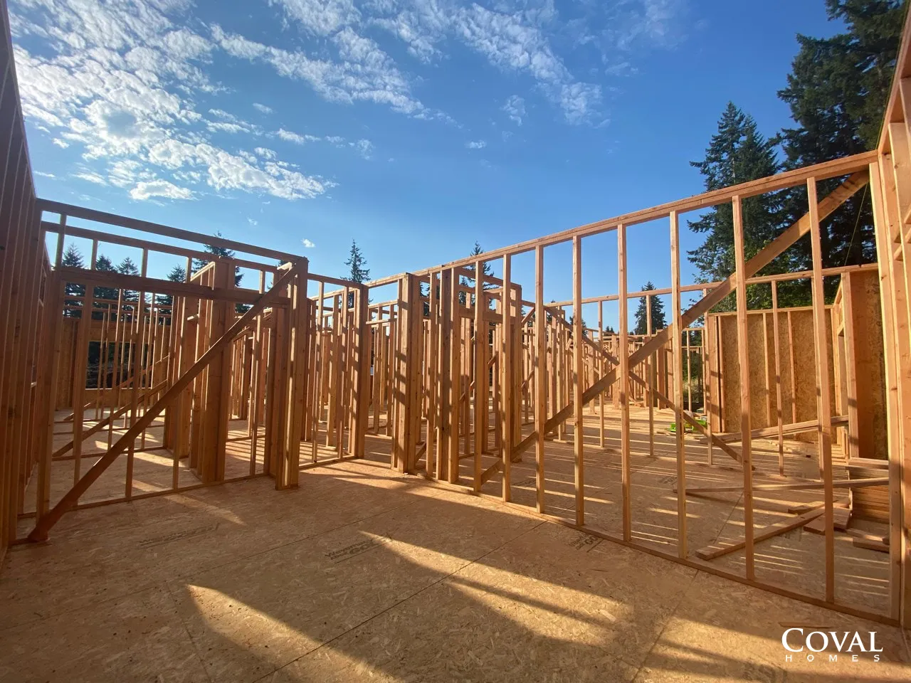 Wood-framed house under construction with exposed walls and roof trusses against a blue sky
