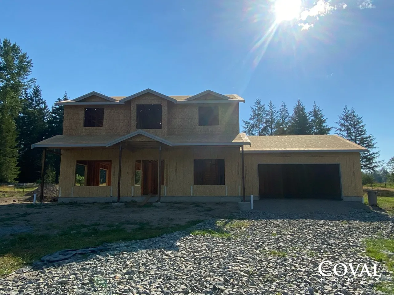 Two-story unfinished house with a stone driveway, garage, and bright sun over a wooded lot