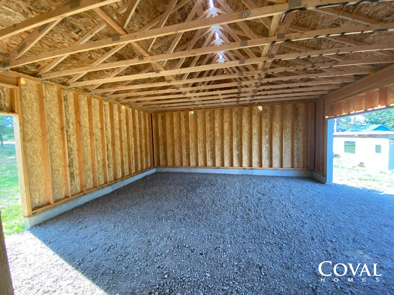 Unfinished wooden garage interior with exposed trusses, insulation walls, and gravel floor openings to the outside