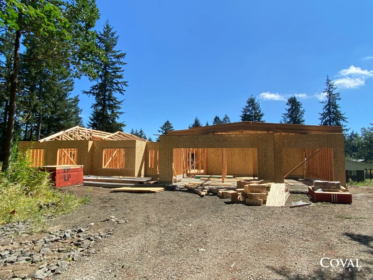 Two unfinished wooden houses under construction on a gravel lot, with trees and a blue sky.