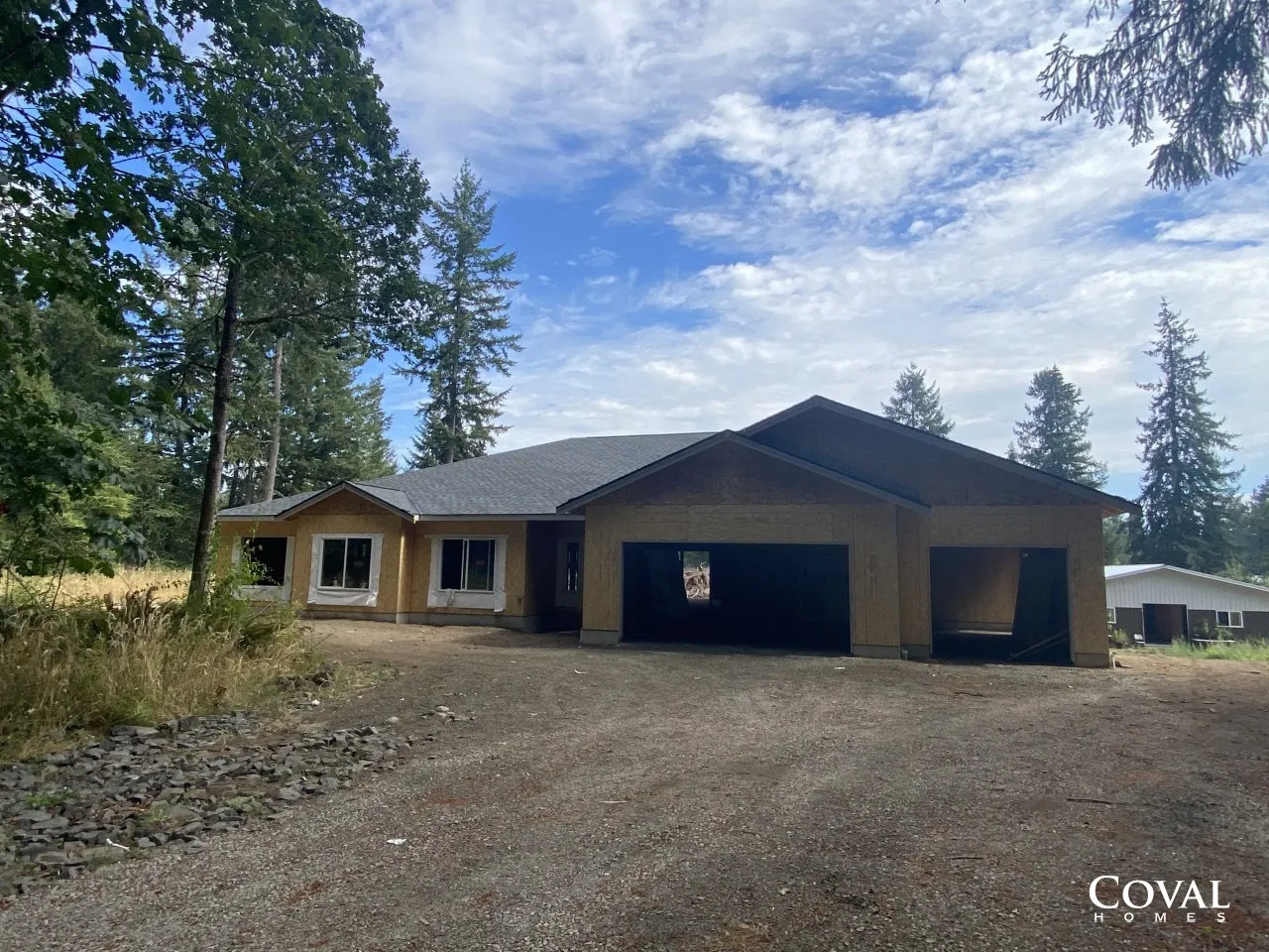 Newly built single-story house with a gravel driveway and surrounding trees under a partly cloudy sky