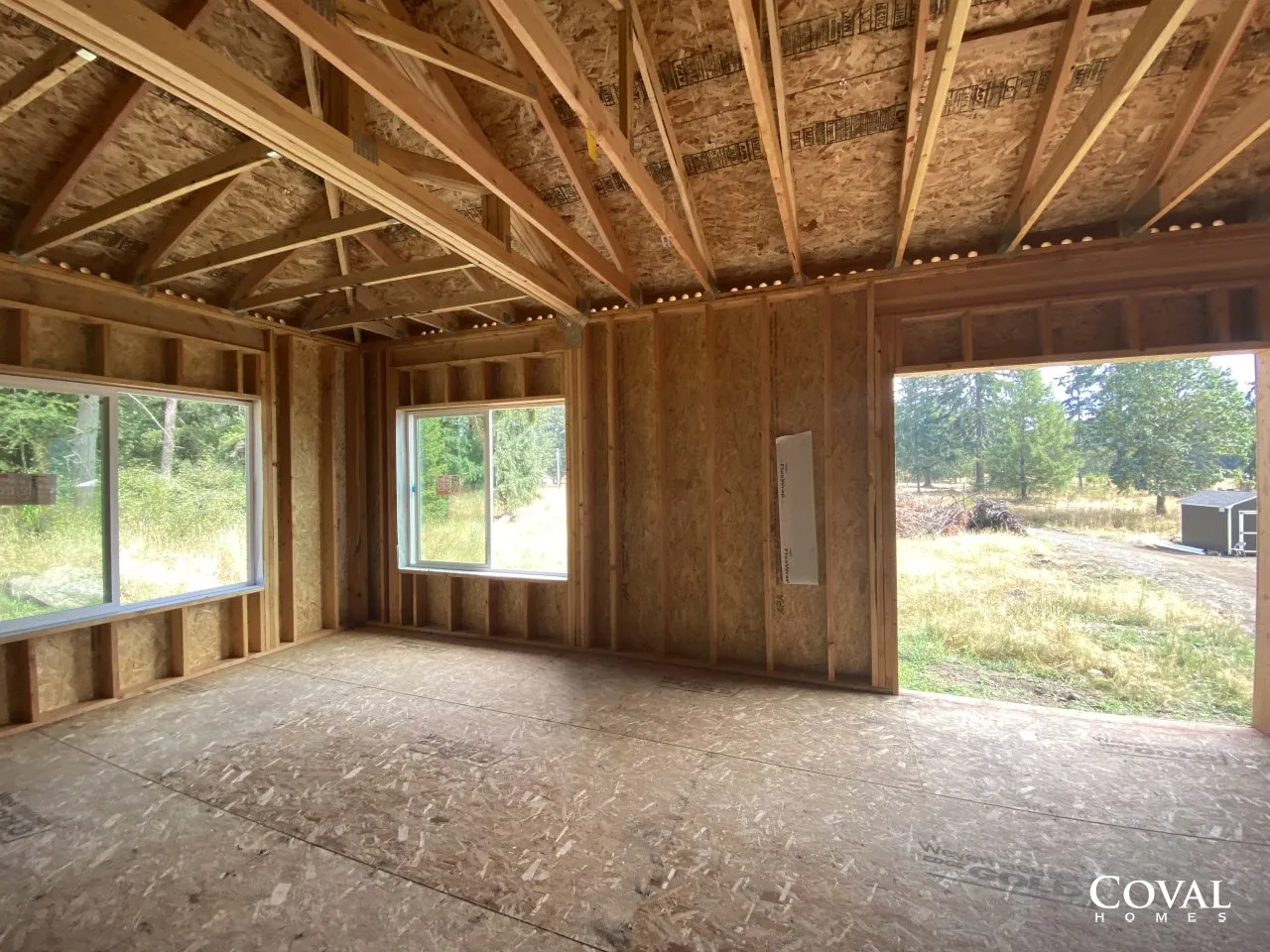 Unfinished wooden room interior with exposed roof trusses, two windows, and a doorway to a grassy yard.