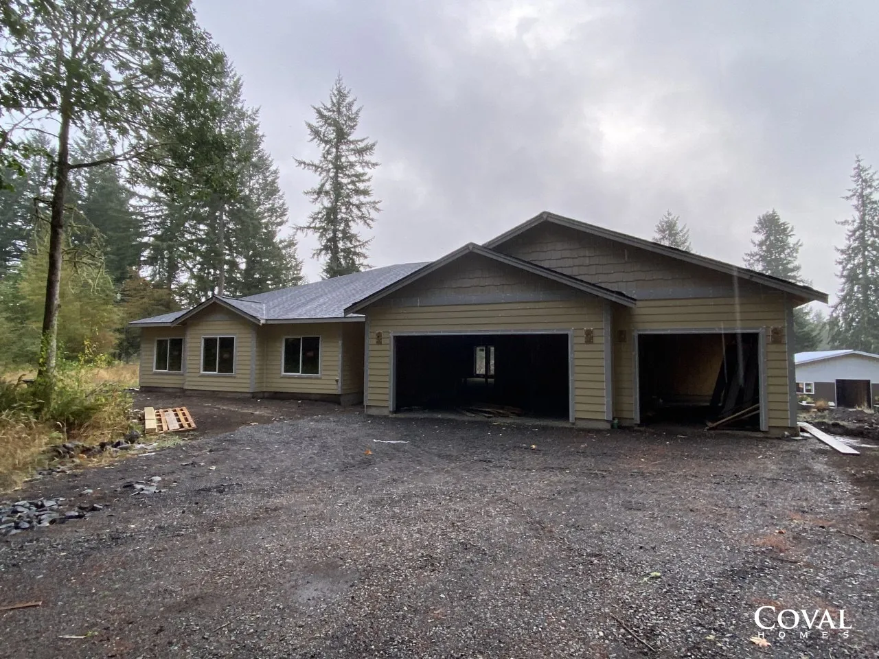 New beige house under construction with attached garage and gravel driveway in a wooded area