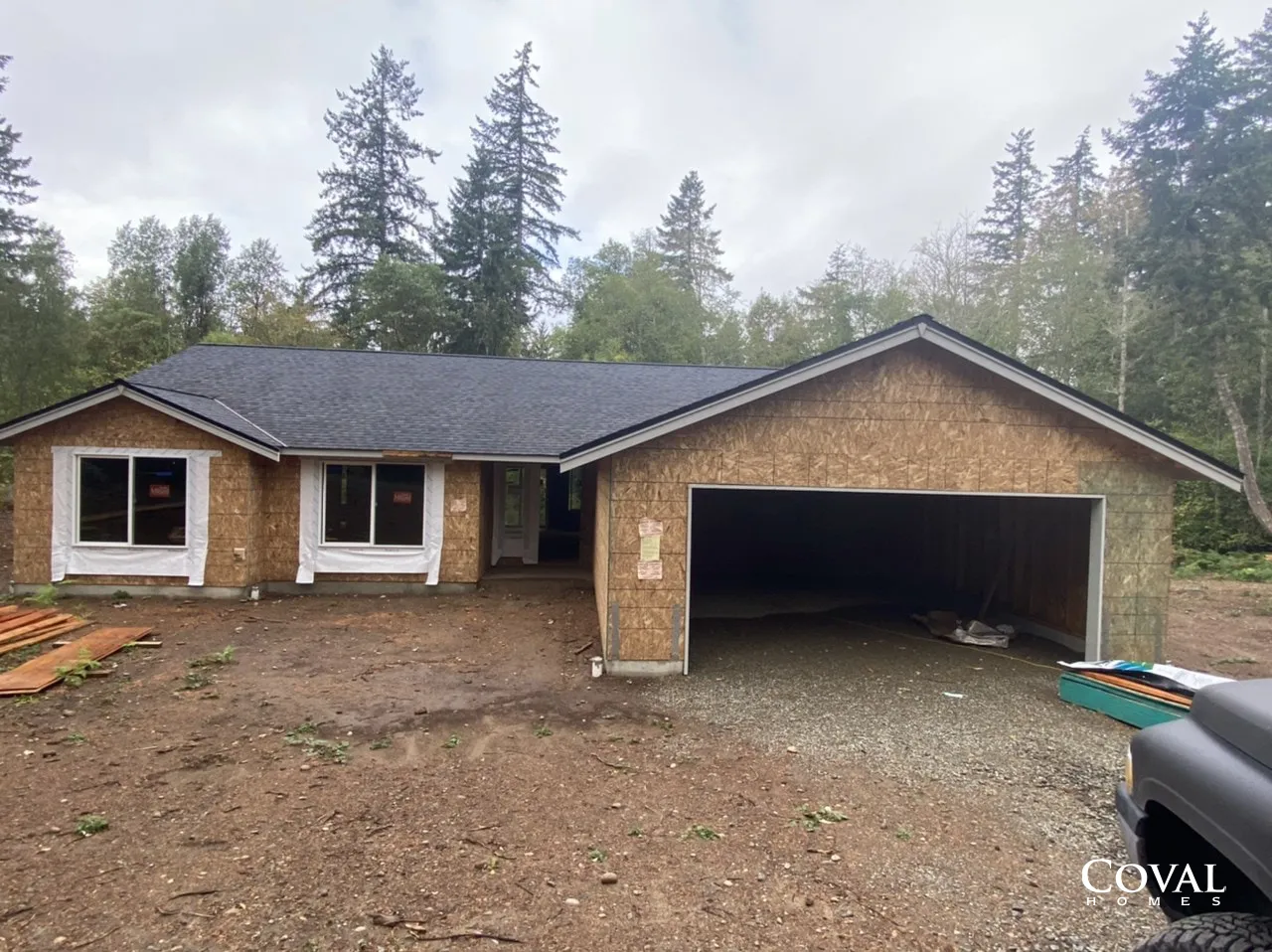 Single-story house with attached two-car garage in a wooded area, under construction on a dirt lot.
