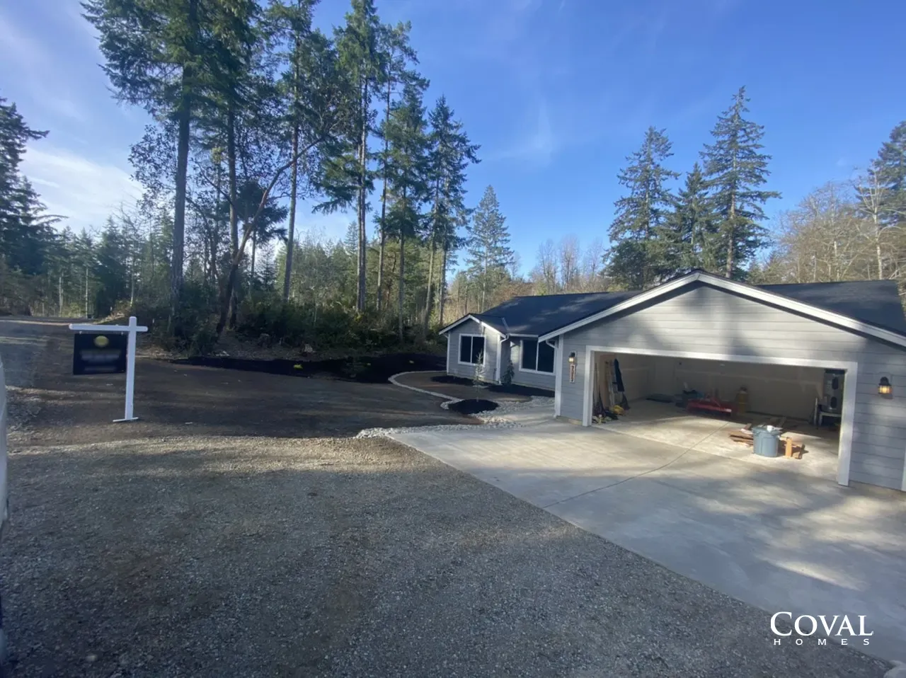Detached gray garage and driveway on a wooded property under a clear blue sky
