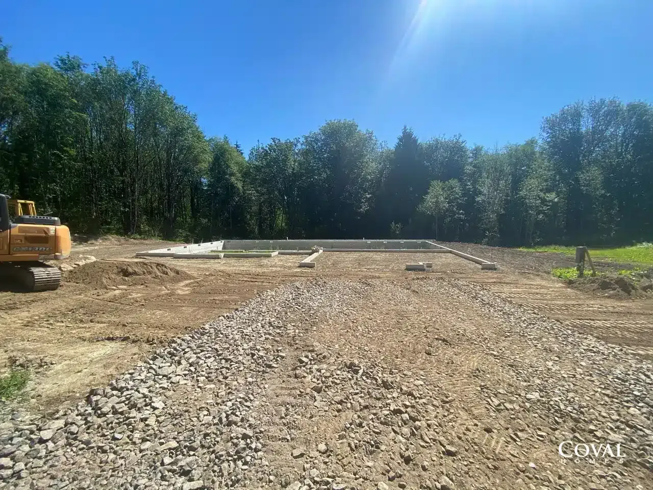 Gravel construction site under a sunny blue sky, with trees along the edge and a bulldozer on the left.
