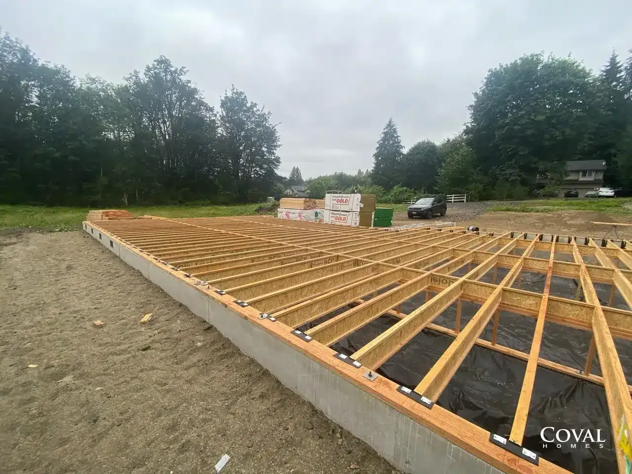 Wooden deck frame under construction beside a gravel lot with trees in the background