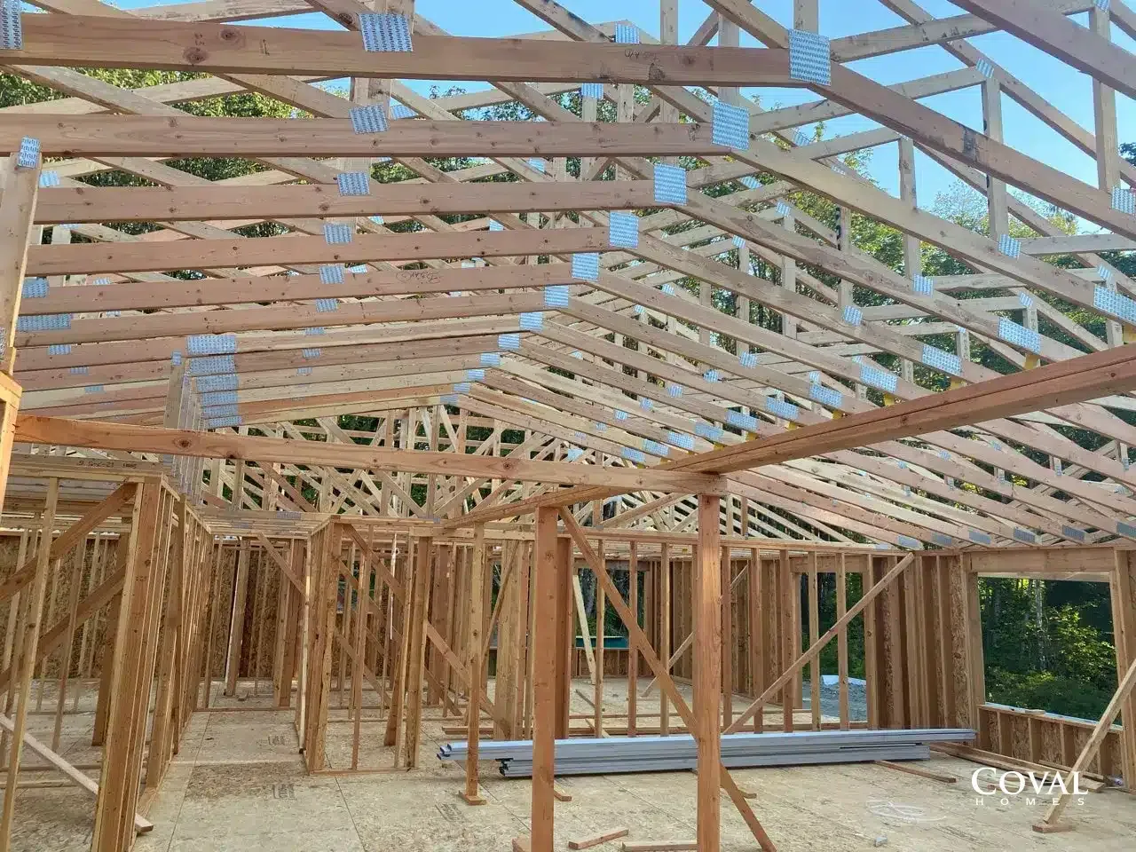 Wooden house framing under construction with exposed roof trusses and walls on a concrete slab.