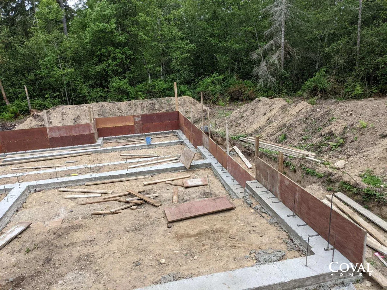 Construction foundation with concrete walls and rebar beside a wooded dirt slope